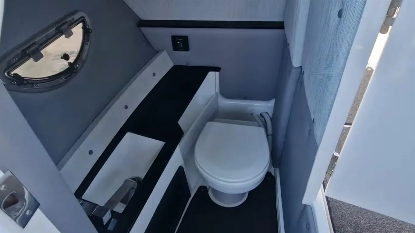 Compact boat bathroom with a toilet, sink area, and porthole for natural light, surrounded by grey and white surfaces.